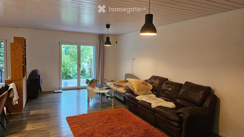 Modern living room with wooden flooring, large sliding glass doors leading to a balcony, a brown leather sofa, a glass coffee table, a small table with a plant, and a hanging pendant light.