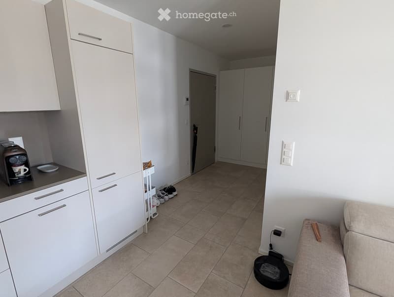 A modern, open-plan living and kitchen area with white cabinets, a coffee maker on the counter, a beige sofa, and several pairs of shoes near the entrance.