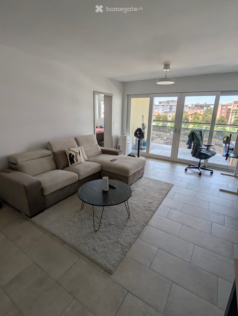Modern living room with a beige sectional sofa, coffee table, and a small round table. The room features large sliding glass doors leading to a balcony with a view of the city.
