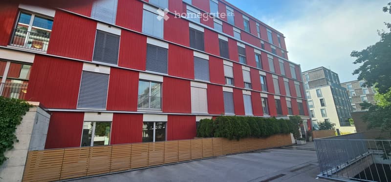 Modern multi-story building with red and grey facade, large windows, and balconies. The ground floor features a wooden fence and greenery.