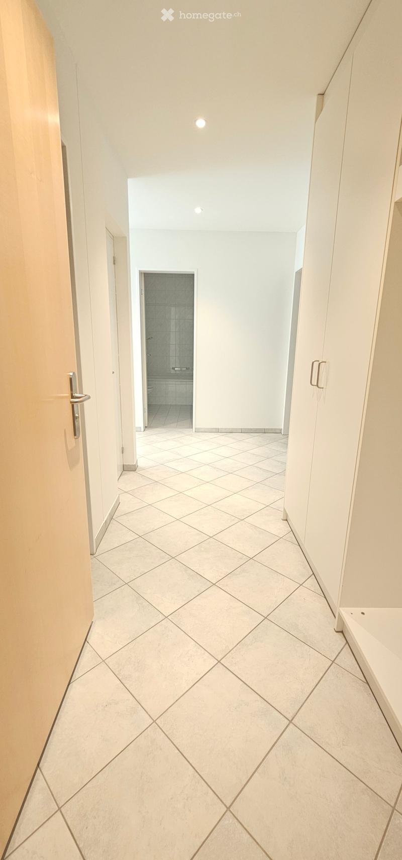Empty hallway with tiled floor, white walls, and a visible bathroom entrance at the end.