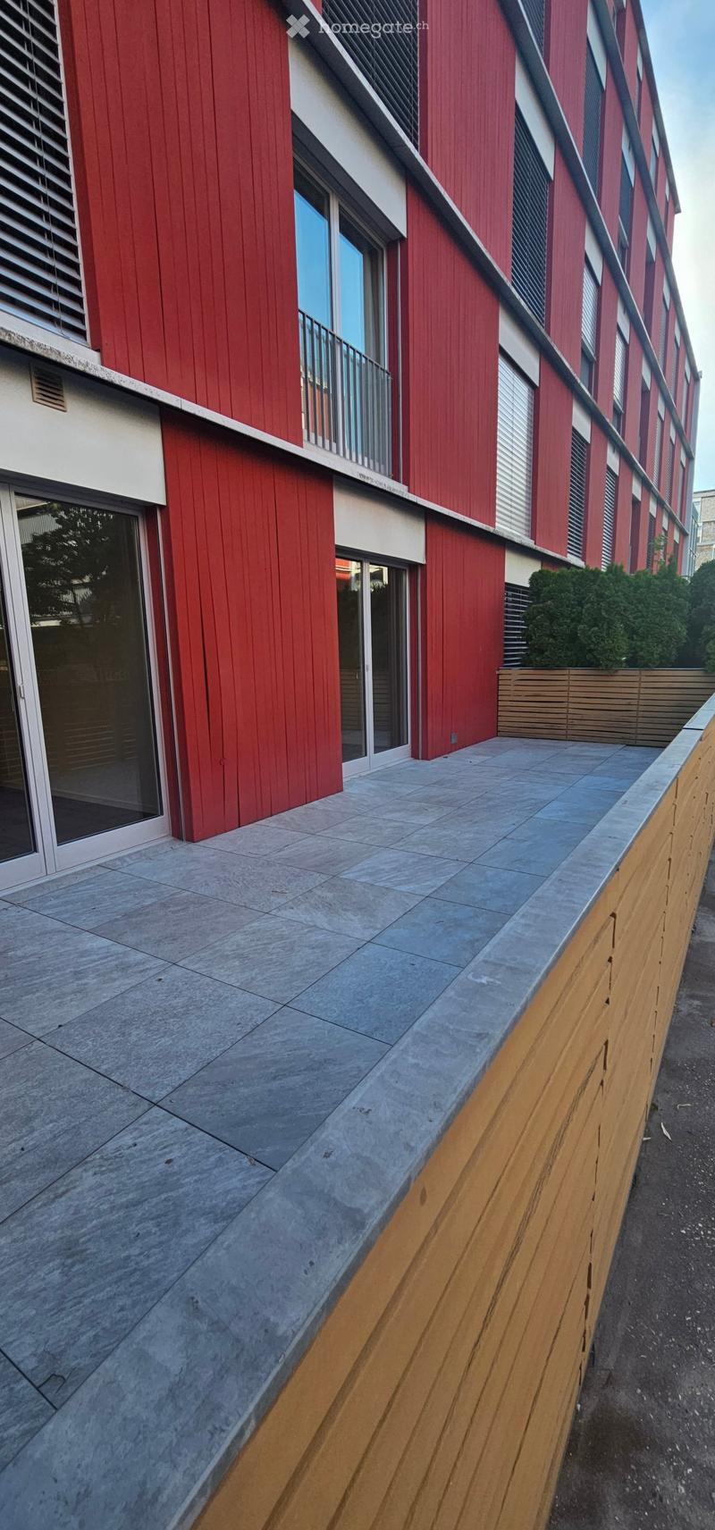 Modern multi-story building with red and white facade, large windows, and balconies. The ground floor features a concrete patio area with wooden decking and a low wooden fence.