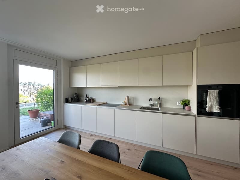 Modern kitchen with white cabinets, wooden floor, and a dining area with a wooden table and chairs. The kitchen features a sink, oven, and various small appliances on the counter.