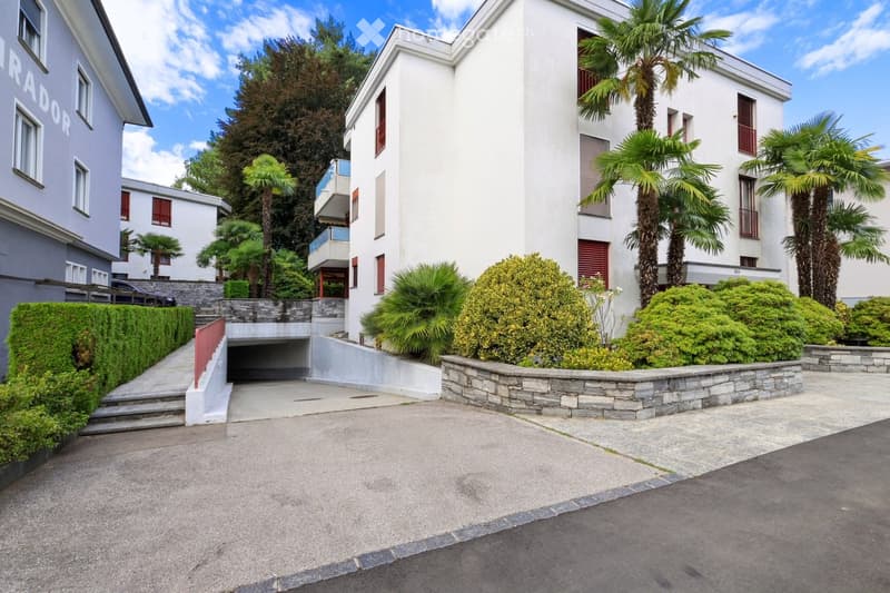 Modern white multi-story building with red shutters, surrounded by palm trees and greenery, with a raised driveway leading to a garage entrance.