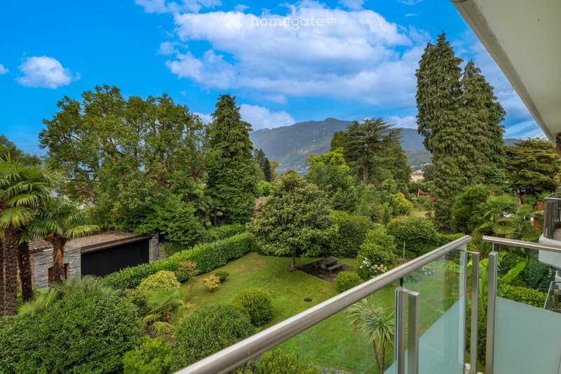 A lush garden with well-maintained greenery, including trees, shrubs, and a small stone structure, with a scenic mountain view in the background.
