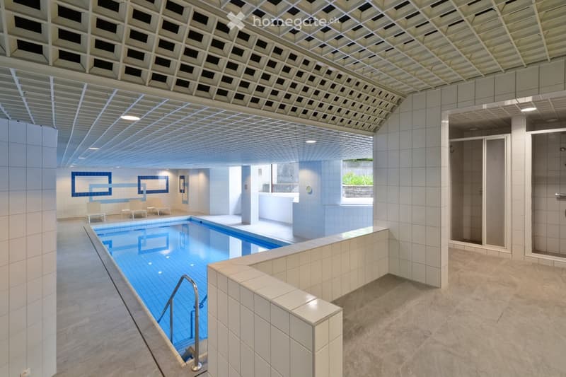 Modern indoor swimming pool area with white tiled walls, a sleek pool, lounge chairs, and a reception desk. The ceiling features a geometric, perforated design with integrated lighting.