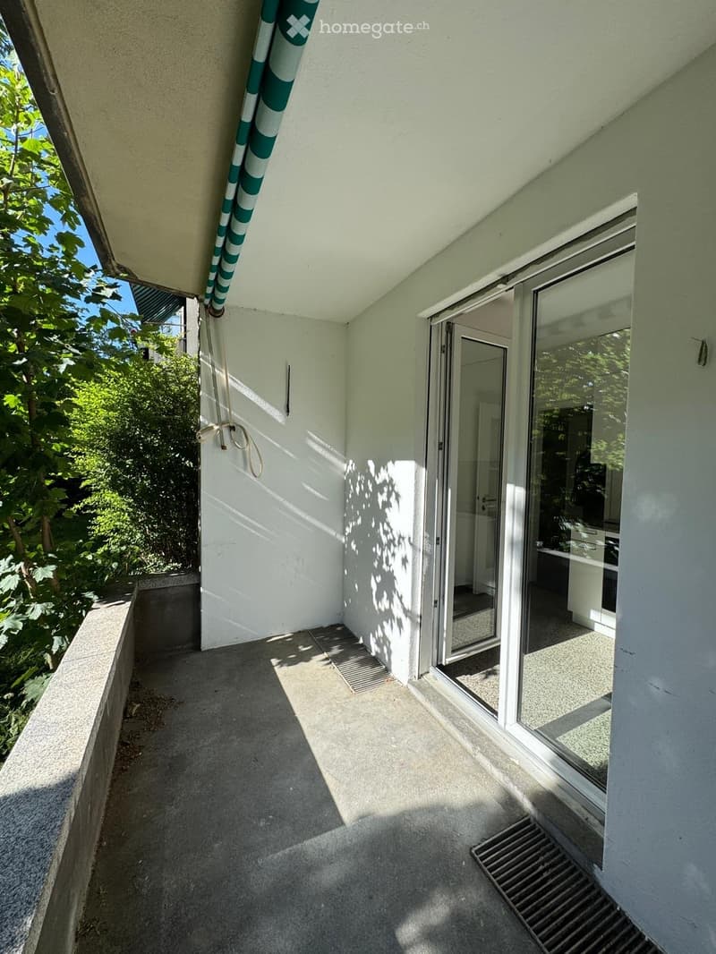 Balcony with concrete floor, glass sliding door leading to interior, green and white striped awning, and a view of greenery outside.