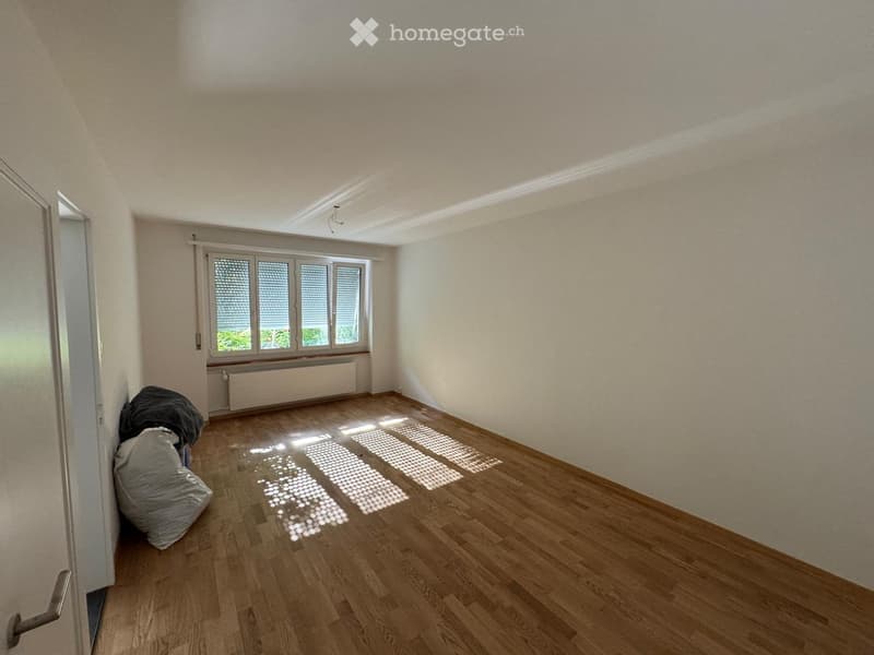 Empty room with wooden flooring, white walls, and a large window with blinds. There are two bags in the corner.