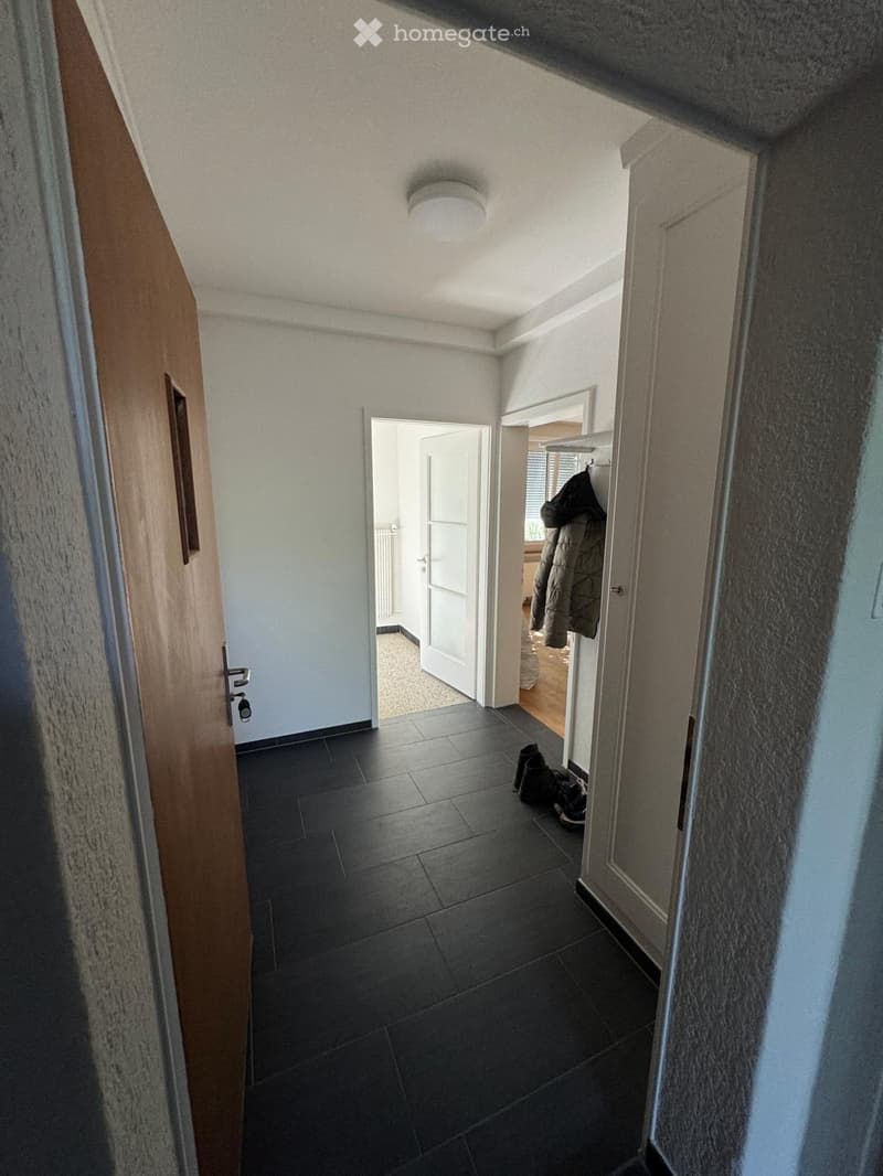A narrow hallway with dark tiled flooring, white walls, and a ceiling light. A coat rack with a jacket and shoes is visible, along with an open door leading to another room.