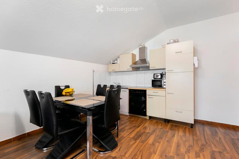 A modern, compact kitchen and dining area with light-colored cabinets, a stainless steel range hood, and a black dining table with six black chairs. The floor is wooden, and the walls are white.