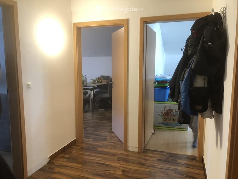 A narrow hallway with wooden flooring and white walls, leading to two rooms. One room contains a dining table and chairs, while the other has a bed and storage items.
