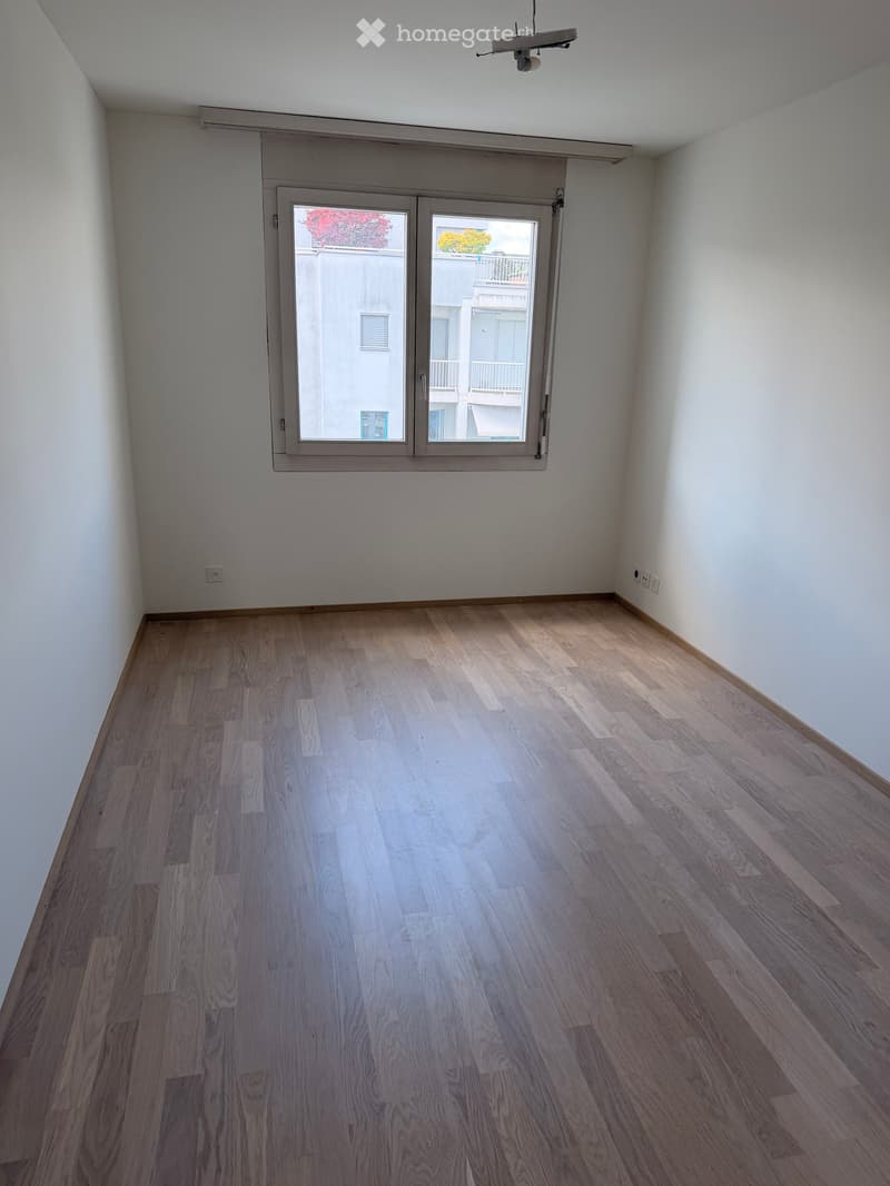 Empty room with light wooden flooring and white walls, featuring a large window with a view of neighboring buildings.