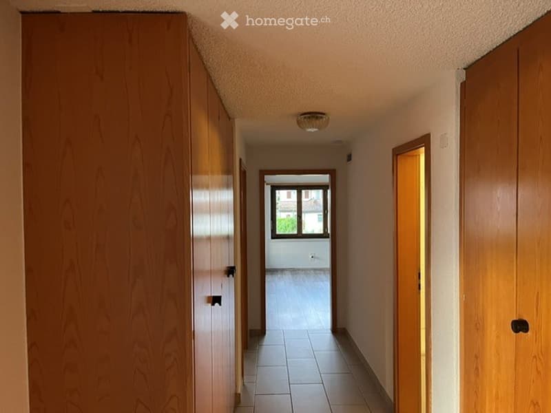 A clean, well-lit hallway with wooden paneling on one side and tiled flooring. A window at the end provides natural light, and there are several doors leading to other rooms.