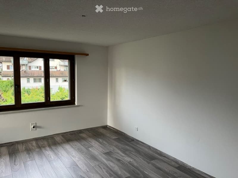 Empty room with wooden flooring, white walls, and a large window with brown frame. No furniture or equipment present.