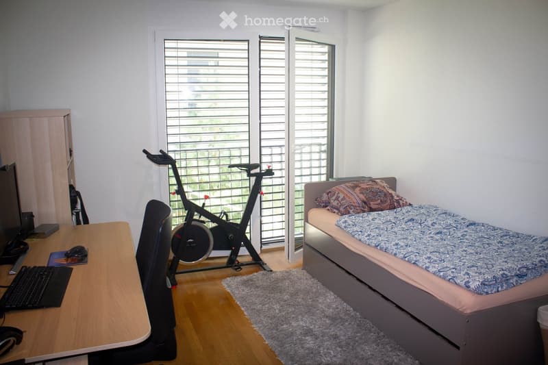 A small, well-lit bedroom with a single bed, desk, exercise bike, and a small rug. The room features large windows with blinds and a wooden floor.