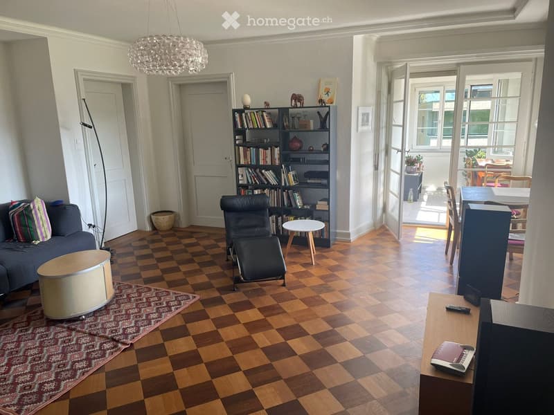 Living room with checkered wooden flooring, a dark blue sofa with colorful pillows, a black leather chair, a small round wooden table, a bookshelf filled with books and decorative items, a television on a wooden stand, and a patterned rug.