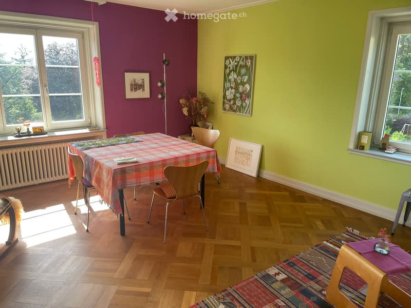 Dining room with wooden floor, colorful walls, table with plaid tablecloth, chairs, picture frames, and a floor lamp.