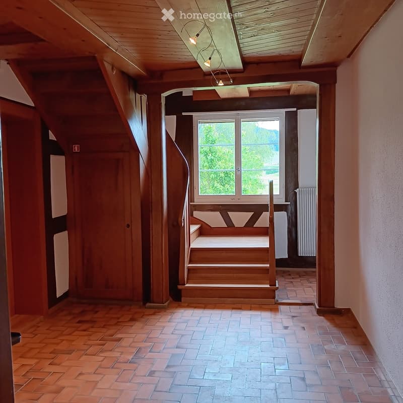 A hallway with wooden beams, a staircase, a window with a view, and a small seating area with wooden benches.