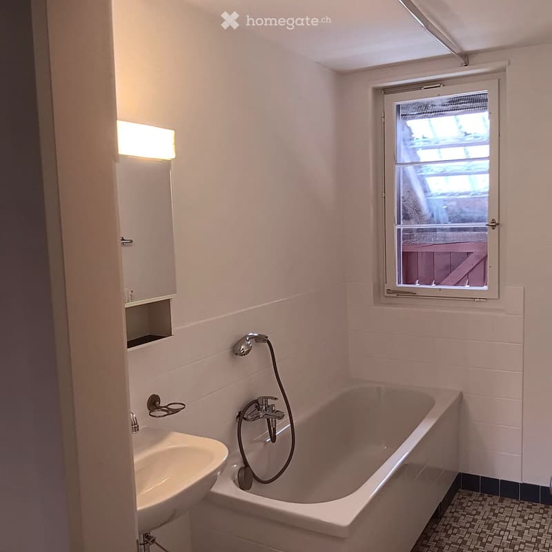 Bathroom with a bathtub, sink, mirror, and light fixture. The room features white tiled walls and a window with a screen.