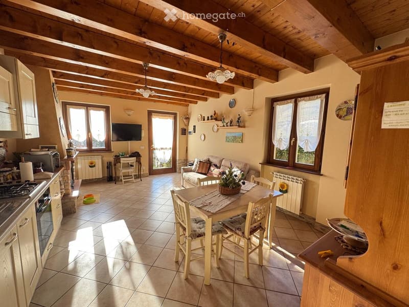 Open-plan living and dining area with exposed wooden beams, tiled floor, and large windows. Features a sofa, dining table with chairs, television, and kitchen appliances.
