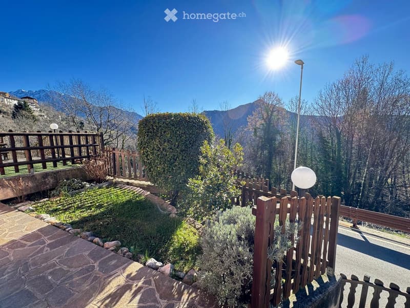 A serene garden area with a wooden fence, neatly trimmed bushes, and a stone pathway, overlooking a scenic mountain backdrop under a clear blue sky.