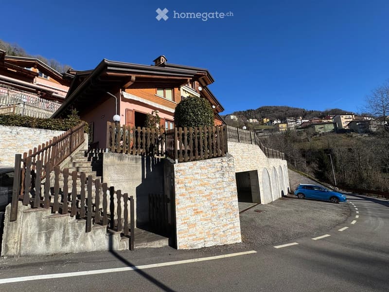Two-story house with a wooden facade, stone staircase, and wooden railing leading to the entrance. A blue car is parked on the street nearby.