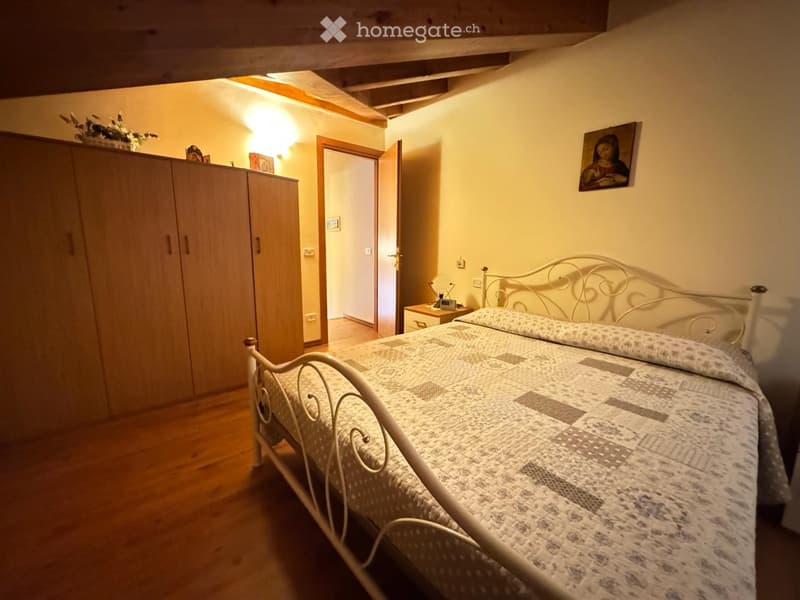 Cozy bedroom with wooden floor, white metal bed, patterned quilt, large wardrobe, nightstand, and artwork on the wall.