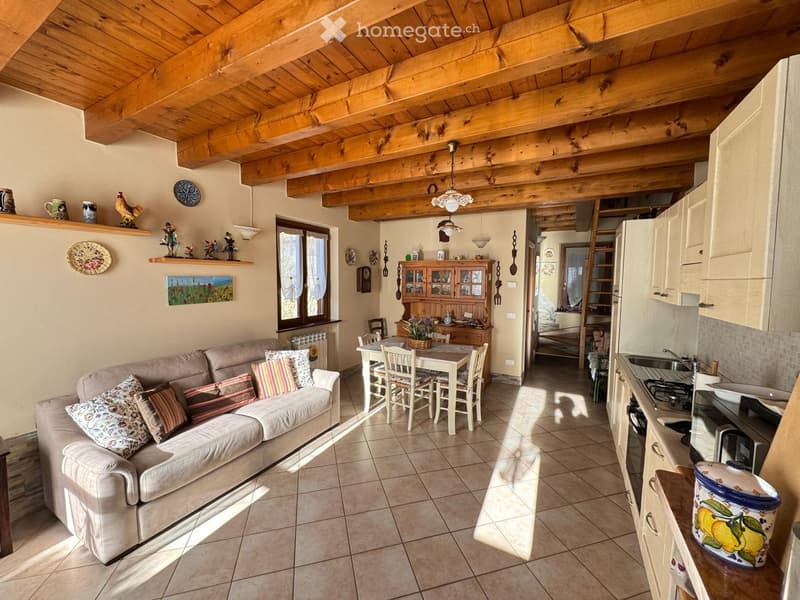 A cozy, rustic living and dining area with exposed wooden beams, a beige sofa with patterned cushions, a dining table with chairs, and a modern kitchen area with white cabinets and stainless steel appliances.
