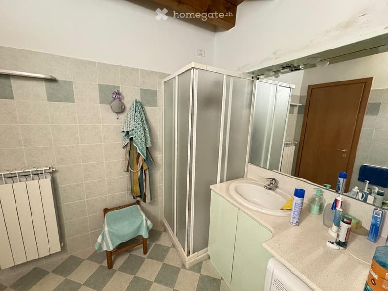 Bathroom with a sink, mirror, shower, radiator, and towels. The sink has various toiletries on the counter, and a small stool is placed near the radiator.