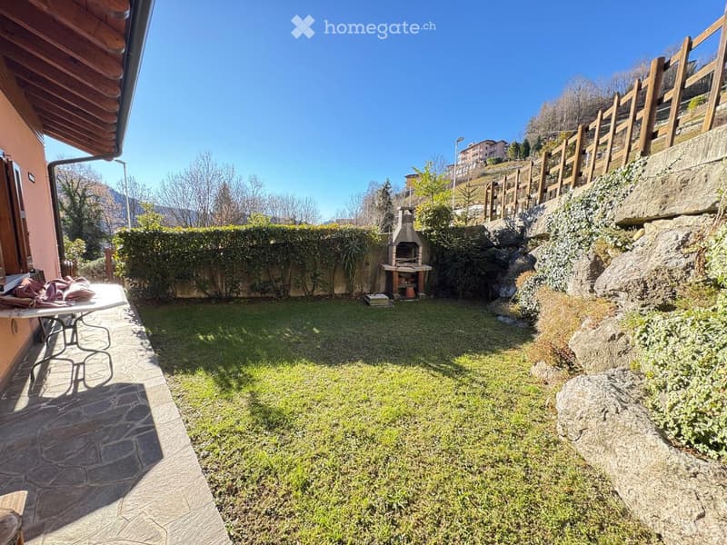 A well-maintained garden with a stone pathway, grassy area, wooden fence, and a small barbecue area. The garden is bordered by a stone wall and has a view of a hillside with trees and buildings in the background.