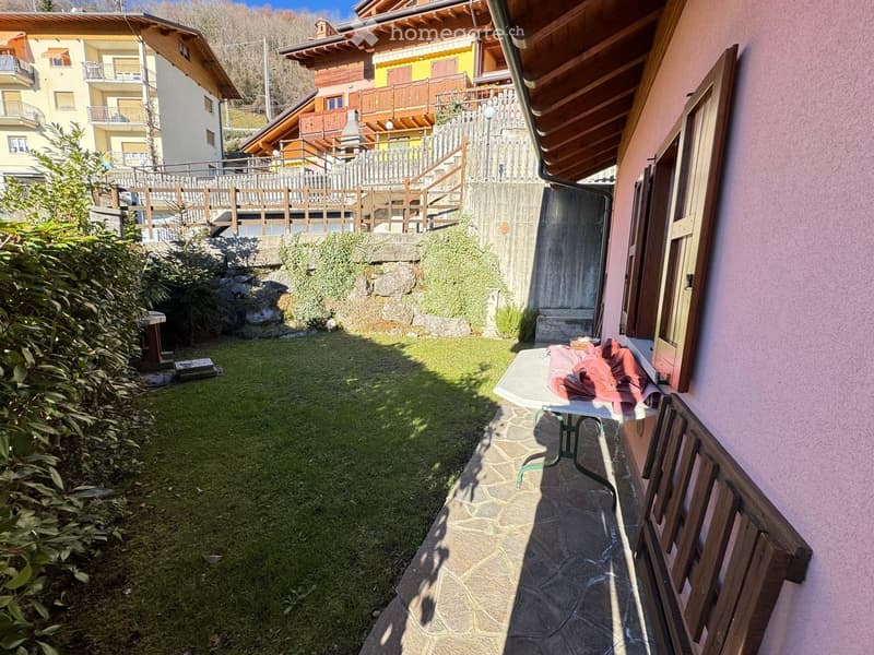 Small garden area with a stone pathway, grassy patch, wooden bench, and a table with folded clothes on it. Wooden fence and stairs lead to a raised wooden deck.