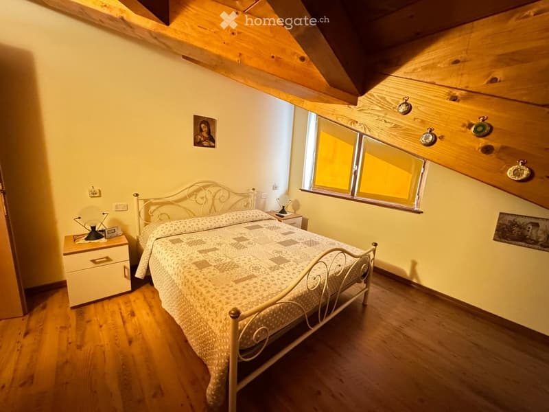 Cozy attic bedroom with wooden slanted ceilings, a metal-framed bed with patterned bedding, a nightstand with a lamp, and a window letting in natural light.