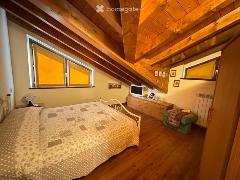 Cozy attic bedroom with wooden slanted ceilings, large windows, a bed with patterned bedding, a nightstand with a lamp, a dresser with a TV, and a small green stool with a blanket.