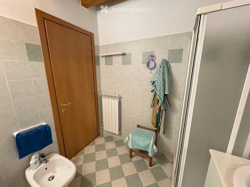 Bathroom with tiled walls and floor, a sink, a radiator, a mirror, and a small stool with towels.