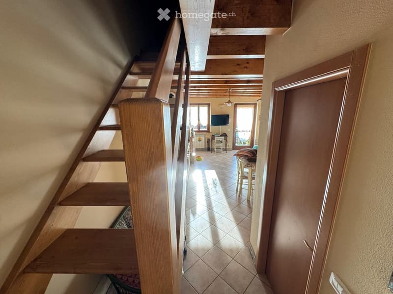 A narrow hallway with wooden stairs leading upwards, beige walls, and a view into a living area with a TV, chairs, and a table.