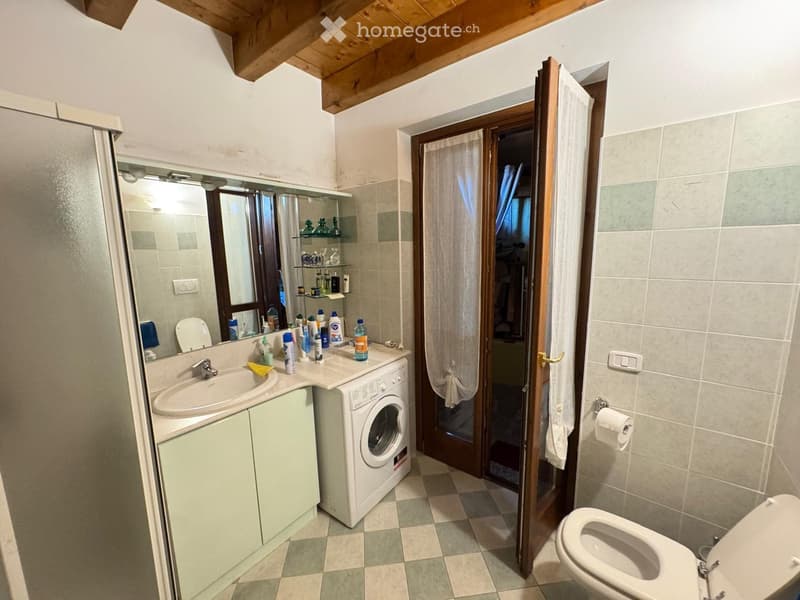 Bathroom with a sink, mirror, toilet, washing machine, and shelves. The room features tiled walls and a wooden door leading to another space.
