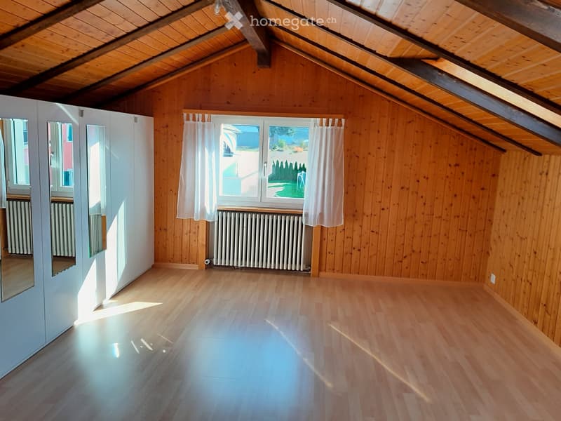 Empty attic room with wooden walls and floor, large window with white curtains, radiator, and built-in wardrobe with mirrored doors.