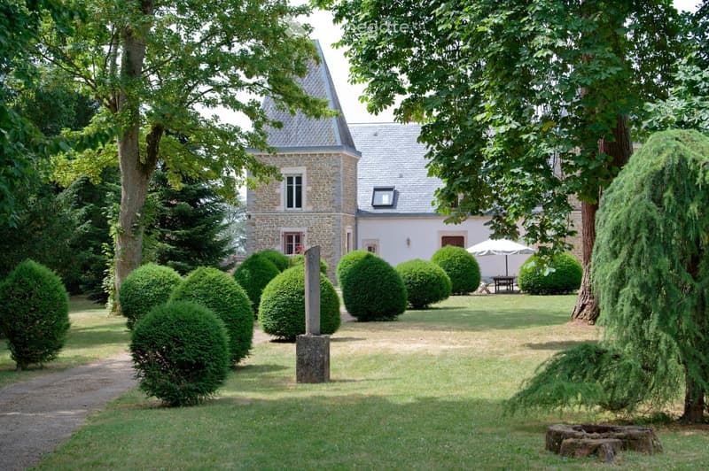 A well-maintained garden with neatly trimmed hedges, a gravel path, and a stone pillar. The background features a two-story house with a grey roof and stone walls, surrounded by tall trees.