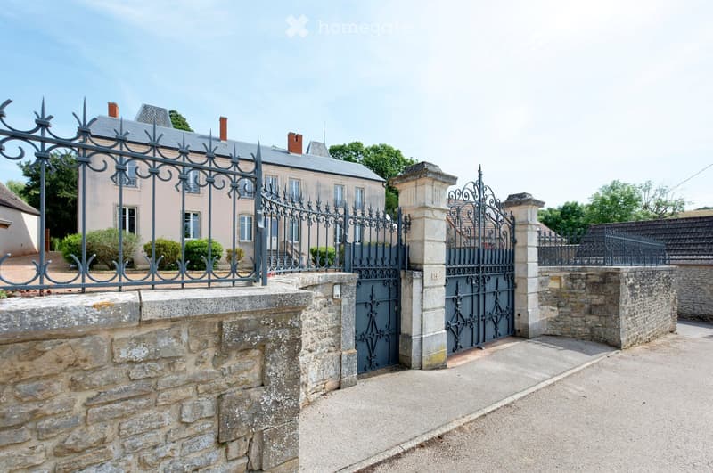 A large, elegant house with a grey roof and light-colored walls, surrounded by a stone wall and ornate metal gates.