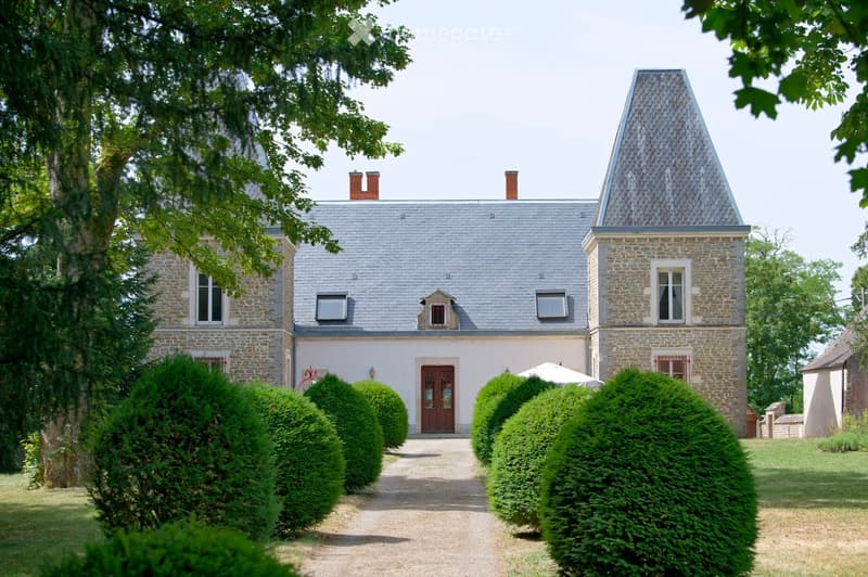A two-story stone house with a grey slate roof, multiple chimneys, and a central entrance with a wooden door. The house is surrounded by well-maintained hedges and trees, with a gravel pathway leading to the entrance.
