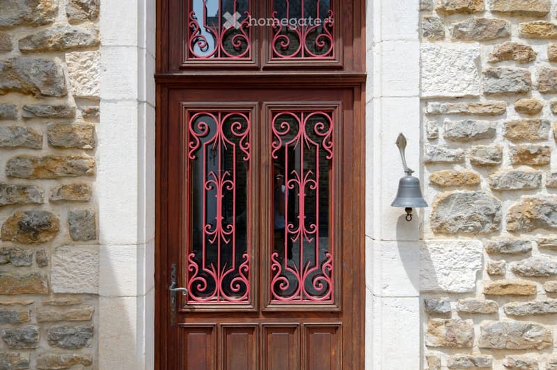 A wooden door with red wrought iron decorative glass panels, flanked by stone walls and a mounted silver bell.