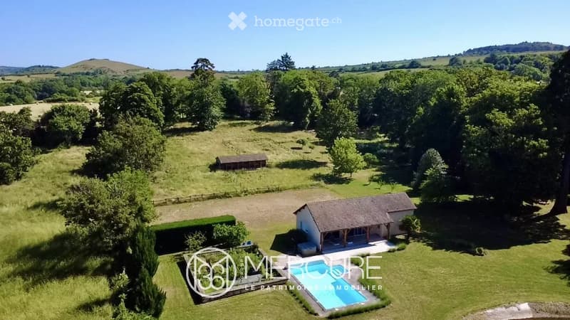 Aerial view of a rural property with a swimming pool, a small house, a barn, and expansive green fields surrounded by trees.