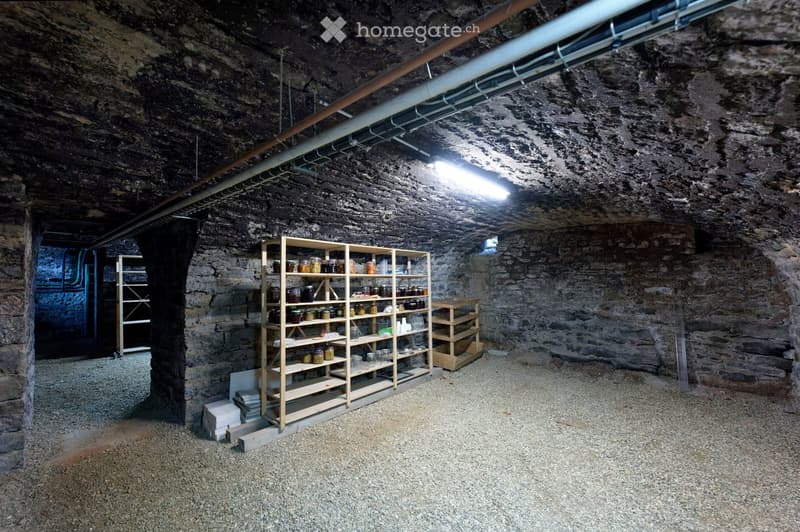 A rustic stone-walled basement with wooden shelves filled with jars and containers, a gravel floor, and exposed pipes on the ceiling.