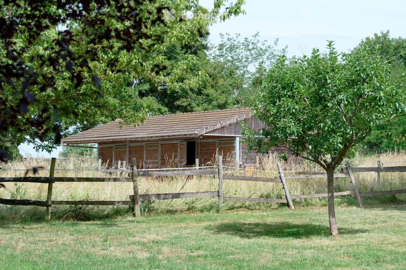 A wooden stable with a tiled roof, surrounded by a wooden fence and trees in a grassy area.