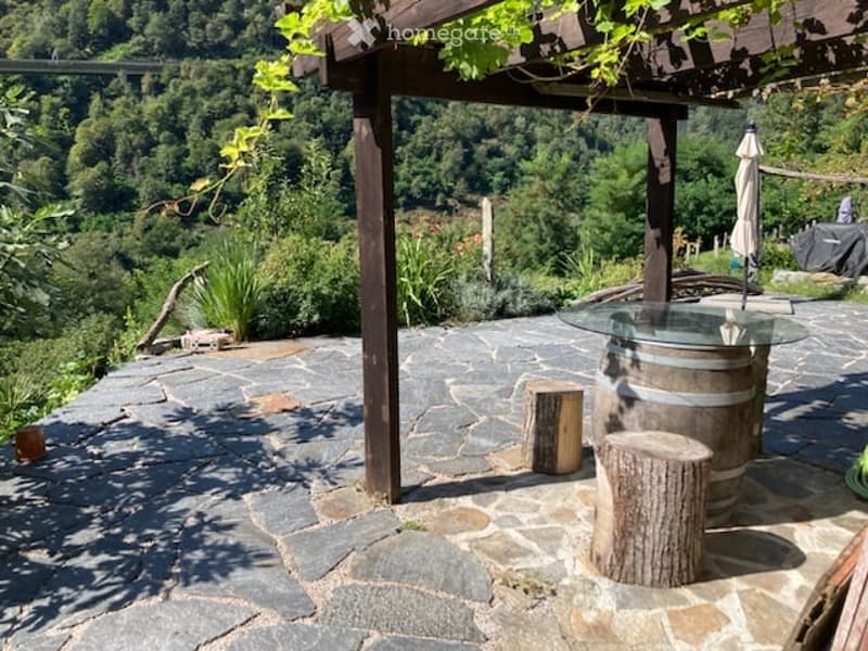 Outdoor patio with stone flooring, wooden pergola, glass table, wooden stools, and scenic mountain view.