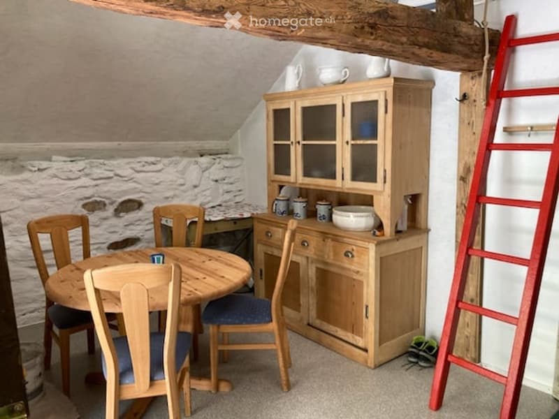 A cozy dining area with a round wooden table surrounded by chairs, a wooden cupboard with glass doors, and various ceramic items on display.