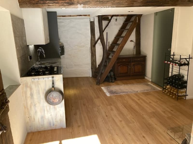 A cozy, rustic kitchen with wooden floors, a small stove, sink, and a wine rack. A wooden staircase leads upwards, and a small rug is placed near the stairs.