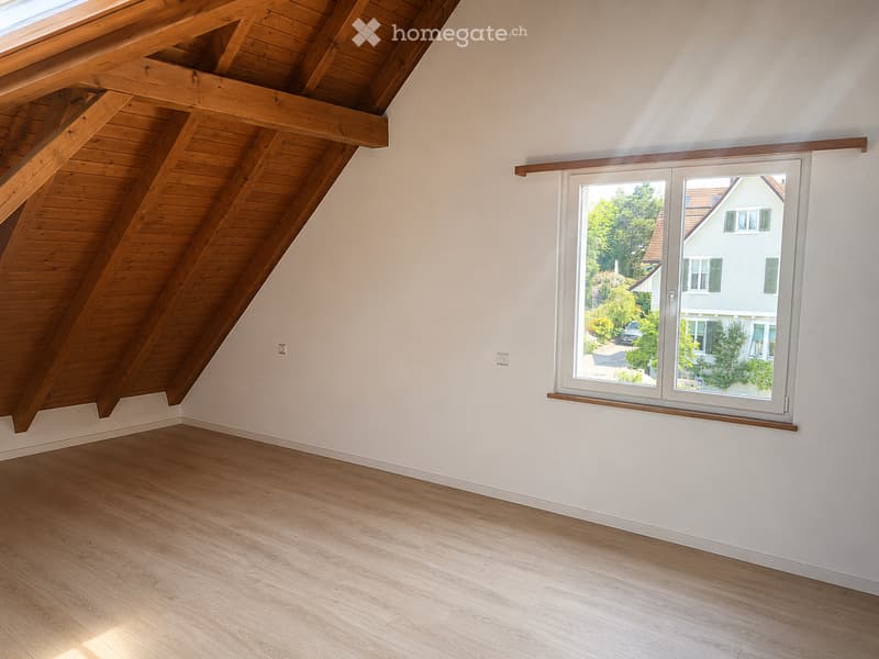 Unfurnished Room. Empty room with wooden floor, white walls, exposed wooden beams on the ceiling, and a large window showing a view of a neighboring house and greenery.