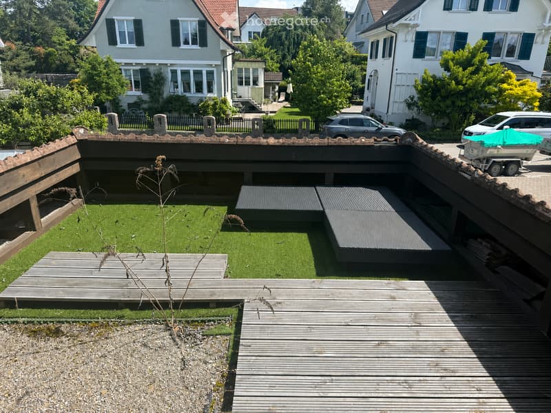 Garden / Outdoor Area. A rooftop garden with artificial grass, wooden decking, and seating areas, surrounded by wooden railings and overlooking a residential neighborhood with houses and trees.