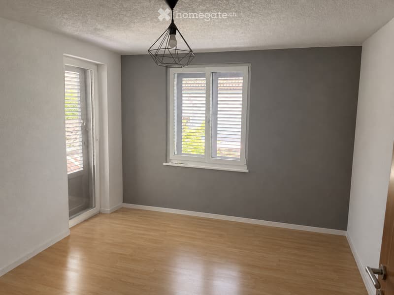 Unfurnished Room. Empty room with wooden flooring, white walls, and a dark accent wall. Features a large window with blinds and a hanging light fixture.